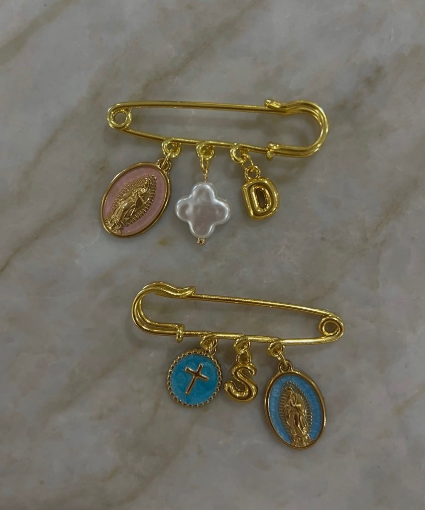 Two gold baby pins: one with a pink Our Lady of Guadalupe medal, a pearl clover, and the letter D; one with a blue cross, the letter S, and a blue Guadalupe medal.